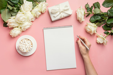 Female hands writing in notebook and bouquet white roses on pink. Top view with copy space.