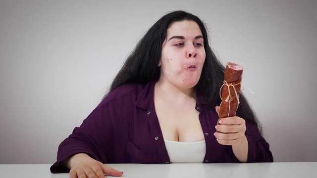 Portrait of plump Caucasian woman smelling and greedily eating sausage. Fat brunette girl hungrily biting food. Obesity, overweight problem, dieting.