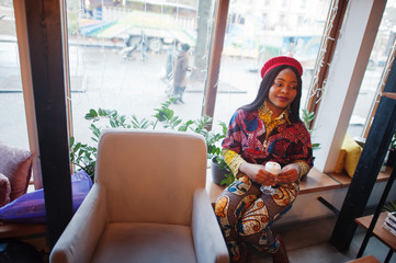 Enthusiastic african american woman in trendy coloured outfit with red beret chilling in cozy cafe with cup of cappucino at hands.