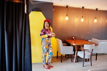 Enthusiastic african american woman in trendy coloured outfit with red beret chilling in cozy cafe with cup of hot latte in hands.