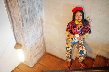 Enthusiastic african american woman in trendy coloured outfit with red beret chilling in cozy cafe, standing on stairs with cup of hot drink in hands.