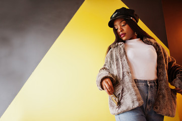 Fashionable african american woman in black cap chilling in cozy cafe and fur against yellow wall.