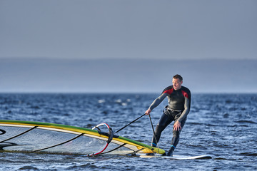 A male athlete is interested in windsurfing. He moves on a Sailboard on a large lake on an autumn day.