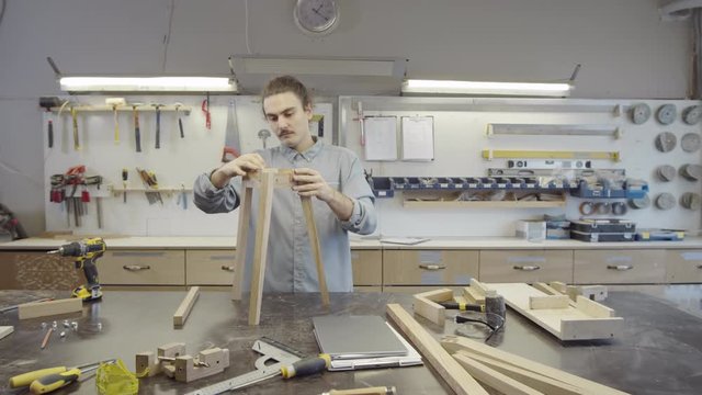 Dollying out waist-up shot of Caucasian carpenter adjusting small wooden components in slots on three-legged base of table while assembling it in workshop, and selection of tools visible in background