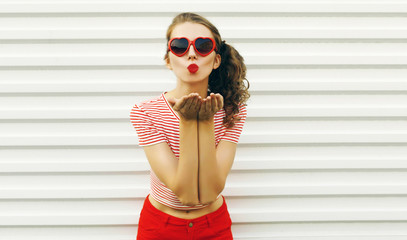 Beautiful young woman blowing red lips sending sweet air kiss wearing red heart shaped sunglasses on white wall background