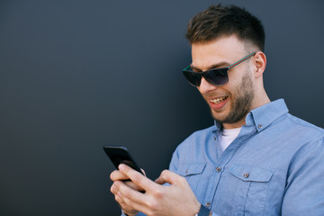 Young man standing against blue wall looking at screen of smartphone and smiling