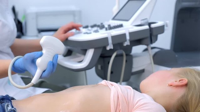 Doctor making abdominal ultrasound for child girl using scanner machine, side view. Woman runs ultrasound sensor over patient tummy looking at screen. Diagnostic examination of internal organs.