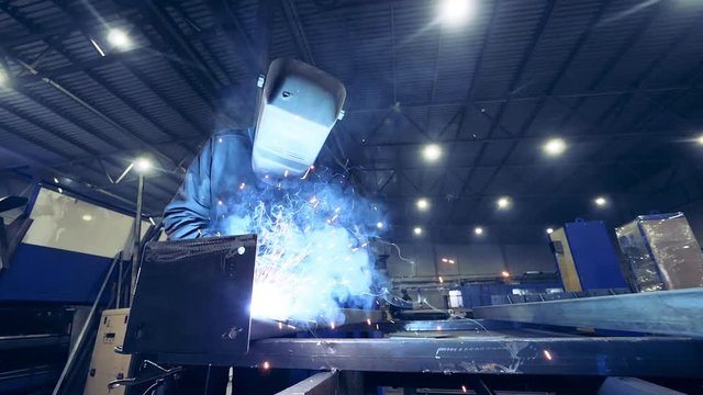 Welder in a helmet is welding iron which causes sparks. Slow motion.