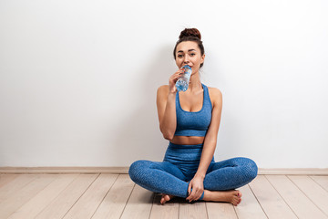 Fototapeta na wymiar The concept of healthy eating. A young white woman in sportswear, with a toned figure, sits on the floor and drinks water from a bottle. White background. Copy space