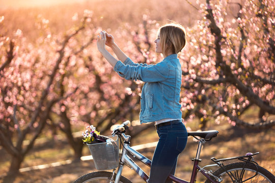 Pretty young woman with a vintage bike taking photographs of cherry blossoms on the field in springtime.