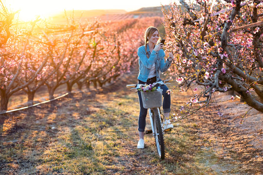 Pretty young woman with a vintage bike taking photographs of cherry blossoms on the field in springtime.