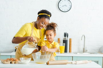 cheerful african american mother pouring milk in bowl near cute daughter in kitchen