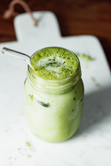 Matcha green tea latte in a glass jar with glass tube on a white marble tray. Healthy clean eating concept.