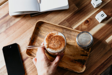 Hand holds a glass of latte served with glass tube on a wooden work desk. Coffee break concept.