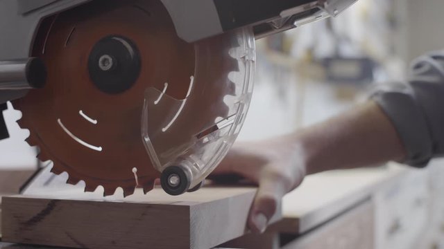 Close-up shot of hands of unrecognizable male woodworking artisan cutting off thin pieces of wood from lumber blank on miter saw at specialist workshop, with accuracy and precision