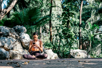 Be alive. Full-length shot of caucasian woman sitting on a mat and using smartphone while going to practice yoga outdoors, in a garden. Healthy lifestyle and relaxation concept. Horizontal shot