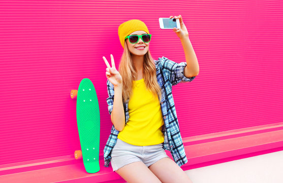 cool smiling woman taking selfie picture by smartphone with skateboard wearing colorful yellow hat on pink background