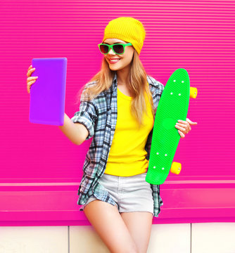 cool smiling woman taking selfie picture by tablet pc with skateboard wearing colorful yellow hat on pink background