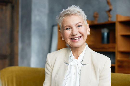 Portrait of experienced mature female professional with short pixie hairstyle and broad confident smile welcoming you in her office wearing elegant beige suit. Age, career and success concept