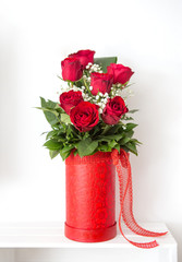 The bouquet of red roses is in a handmade red box. A rose arrangement is a beautiful gift for a loved one. Behind the bouquet is a white background.