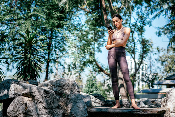 Fototapeta na wymiar Add yoga in your life. Full-length shot of caucasian woman standing and looking at her smartphone while going to practice yoga outdoors, in the nature park. Healthy lifestyle and relaxation concept