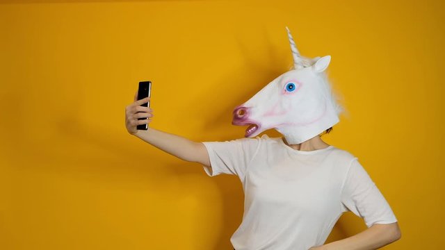 Strange funny video - woman with unicorn head making selfie with smartphone over yellow background