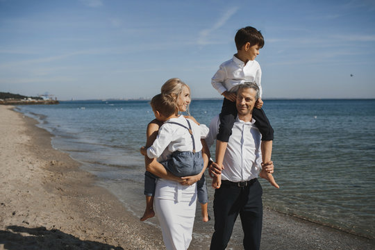 Stylish family, of parents and two sons on their hands, are walking on the sunny summer day on the coastline