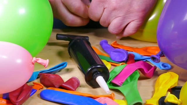 Close POV shot of a man&rsquo;s hands tying a knot in a balloon with many more on a table, plus a small air inflator / pump.