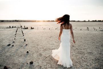 Fototapeta na wymiar Back view of caucasian girl in long white dress without sleeves in the evening on the sandy area