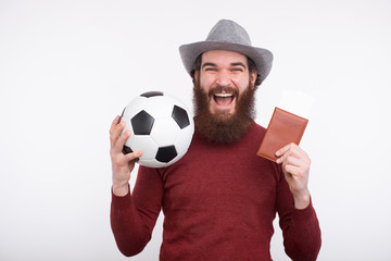Happy bearded man in casual holding soccer ball and passport with tickets