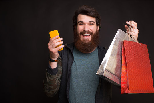 Online shopping is great. Bearded man is holding two shopping bags and his phone on black background.