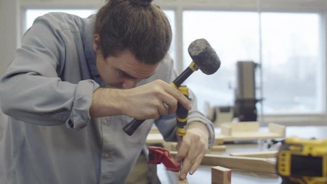 Waist-up shot of Caucasian male carpenter bending over worktop at workshop and carefully cutting small slots in wooden furniture component, hammering chisel with mallet