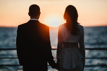 Back view of a romantic couple on the sunset by the sea who are holding hands together