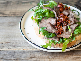 Large toast on homemade bread with soft cheese, arugula and slices of roast beef with sun-dried...