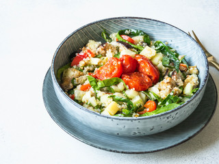 Salad of quinoa, baked vegetables, spinach and raw cucumber in a blue plate on a white wood background