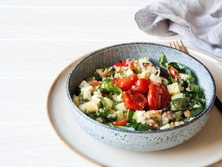 Salad of quinoa, baked vegetables, spinach and raw cucumber in a blue plate on a white wood background
