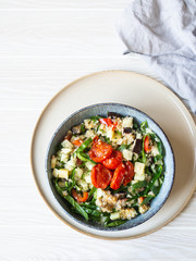 Salad of quinoa, baked vegetables, spinach and raw cucumber in a blue plate on a white wood background