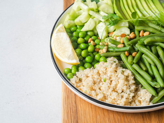 Quinoa bowl with green beans, green peas, cucumber, avocado, and pine nuts on wood cutting board on grey background.