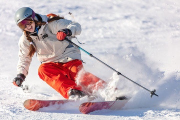 Girl On the Ski. a skier in a bright suit and outfit with long pigtails on her head rides on the track with swirls of fresh snow. Active winter holidays, skiing downhill in sunny day. Woman skier - obrazy, fototapety, plakaty