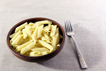 Pasta. Penne in a clay plate on a linen background. Fork on a table.