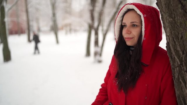Portrait of young woman in red jacket and hood in winter season. Beautiful lady smiles, rejoicing in fall of snow. Fluffy snow envelops everything around.