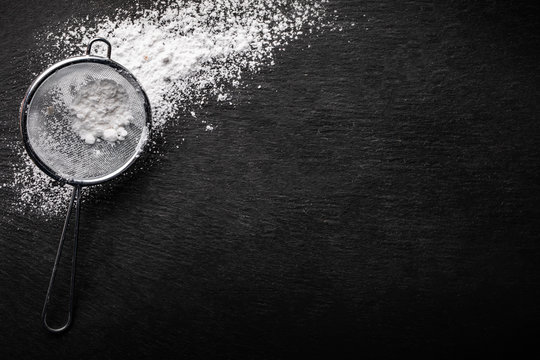 Powdered sugar scattered on a black stone countertop with a sifter for sifting , top view.