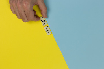 person hand holding domino pieces and play the game on the two-tone yellow and blue surface, top view