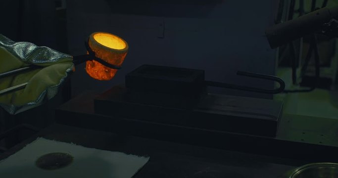 Factory Worker in Silver Gloves Holds the Hot Gold in a Vessel Using Metallic Tongs and Pours Liquid Metal into Shape. Machine Starts the Fire. Gold Bars Production