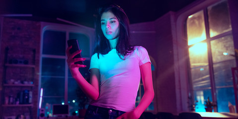 Naklejka na ściany i meble Serfing. Cinematic portrait of handsome stylish woman in neon lighted interior. Toned like cinema effects in purple-blue. Caucasian female model using smartphone in colorful lights indoors. Flyer.