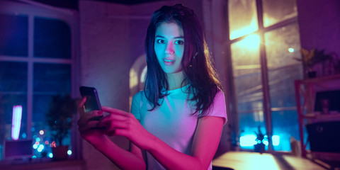 Smiling, looking forward. Cinematic portrait of stylish woman in neon lighted interior. Toned like cinema effects in purple-blue. Caucasian model using smartphone in colorful lights indoors. Flyer.