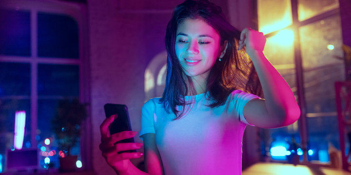 Taking selfie. Cinematic portrait of handsome stylish woman in neon lighted interior. Toned like cinema effects in purple-blue. Caucasian model using smartphone in colorful lights indoors. Flyer.