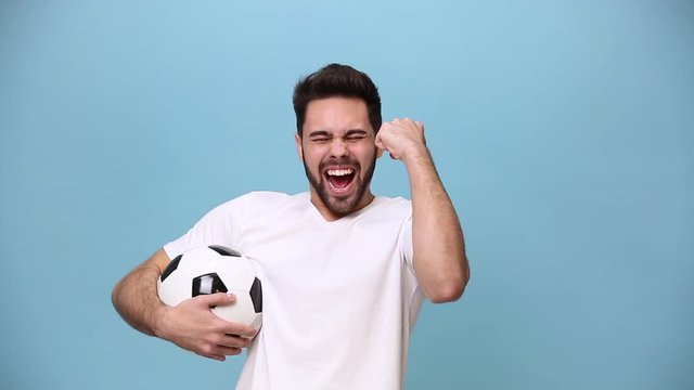 Bearded young guy 20s football fan cheer up support favorite team with soccer ball expressive gesticulating hands, in white t-shirt isolated on pastel blue background in studio. People sport concept