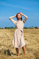 Young beautiful brunette model in a beige dress posing against the background of a mowed wheat field