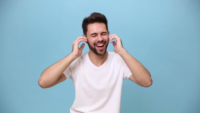Handsome unshaven bearded young guy 20s in white t-shirt isolated on pastel blue background in studio. People sincere emotions lifestyle concept. Listen music in earphones dance chill enjoy hands up